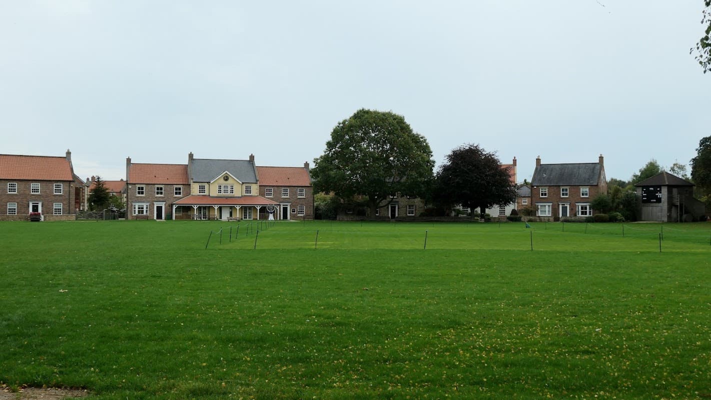 North Stainley Village Hall surrounded by green grass, trees, and nearby buildings under a cloudy sky.
