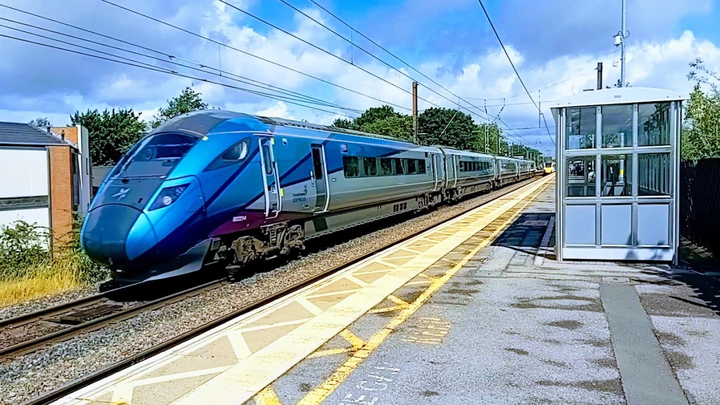 A sleek blue train passes by a bus stop with a glass shelter in Northallerton, under a partly cloudy sky.