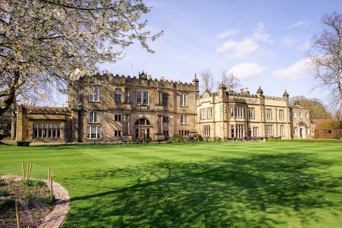 Historic stone building with manicured lawns, surrounded by trees and blooming flowers under a clear blue sky.