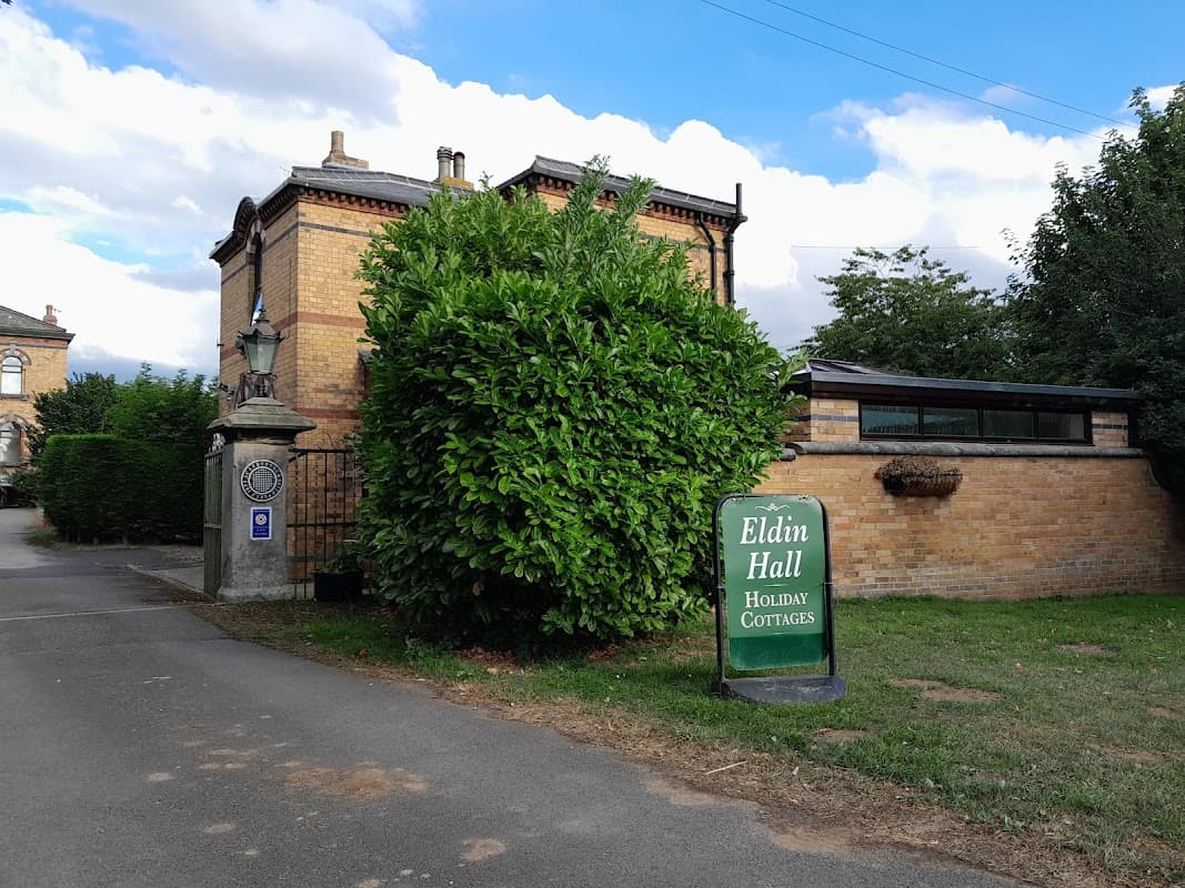 Eldin Hall Cottages sign near a lush green bush and brick building, with a cloudy sky in the background.