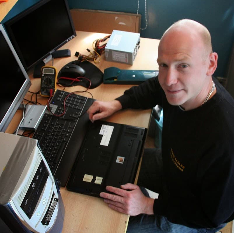 A man working on a laptop at a desk with monitors, tools, and computer components in a tech workspace.