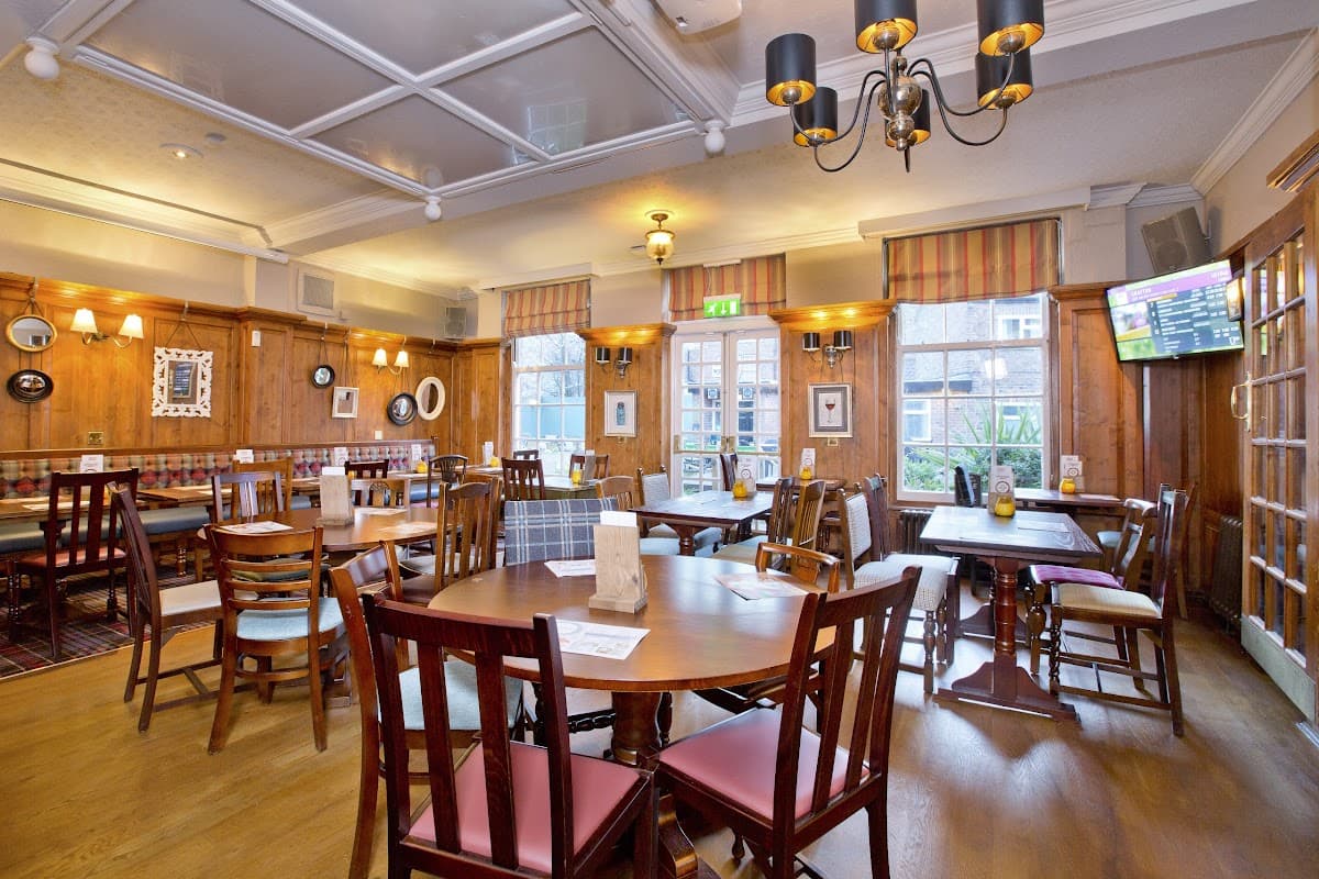 Cozy pub interior with wooden tables, chairs, and warm lighting, featuring large windows and a TV in the corner.