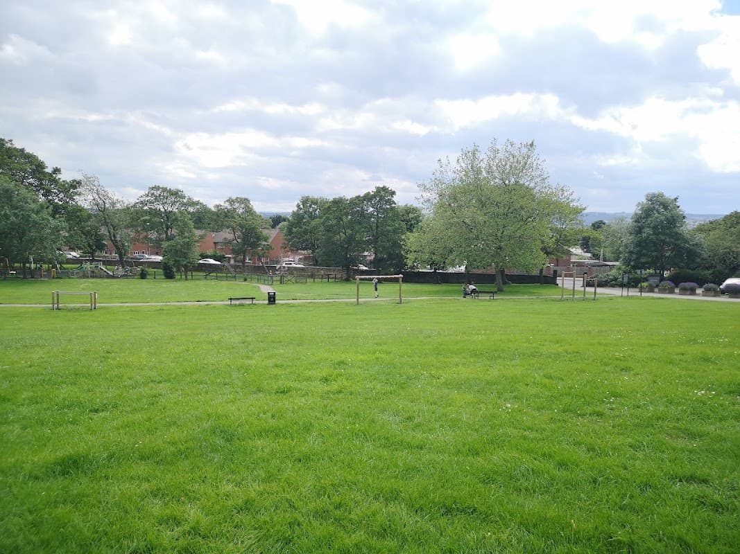 Lush green park with trees, benches, and a playground, set against a cloudy sky in Ossett, Yorkshire.