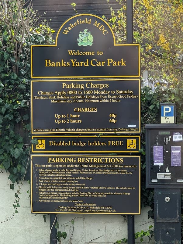 Sign for Banks Yard Car Park in Ossett, detailing parking charges, hours, and restrictions, surrounded by greenery.