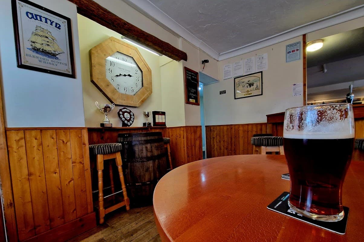 Cozy bar interior with wooden paneling, a large clock, trophies, and a pint of dark beer on a table.