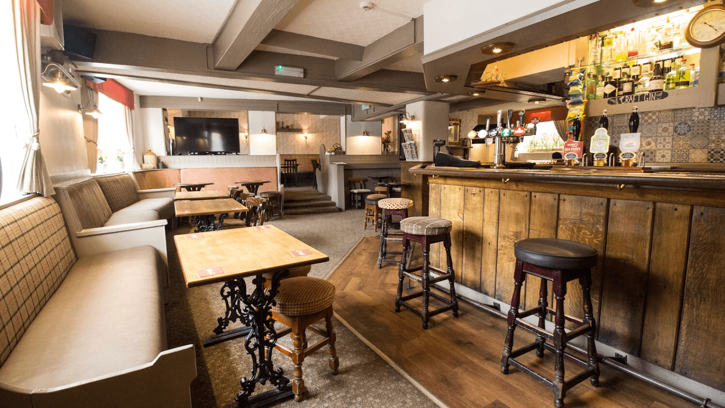 Cozy interior of The Cock Inn with wooden bar, tables, and stools, featuring warm lighting and a welcoming atmosphere.