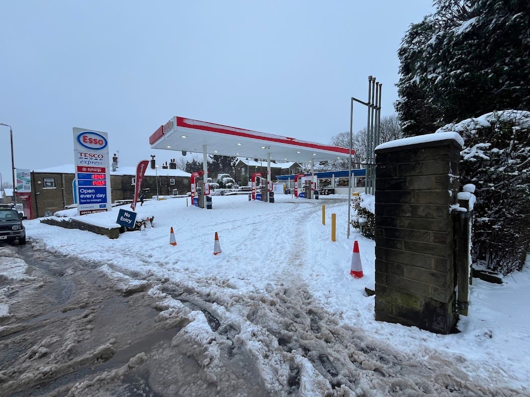 Esso gas station in Ovenden, Yorkshire, surrounded by snow, with visible fuel pumps and a sign indicating open hours.