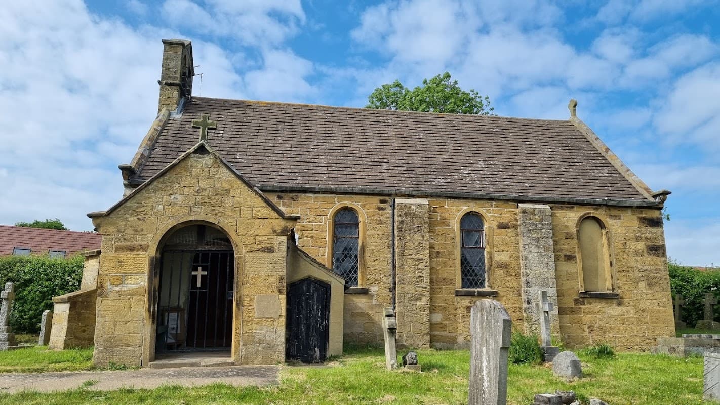 Stone building with a cross on the roof, surrounded by greenery and gravestones under a partly cloudy sky.