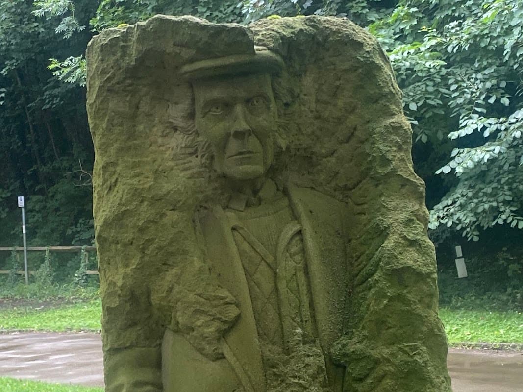 Stone sculpture of a man in a hat, set amidst greenery near Nidd Walk Parking in Pateley Bridge, Yorkshire.