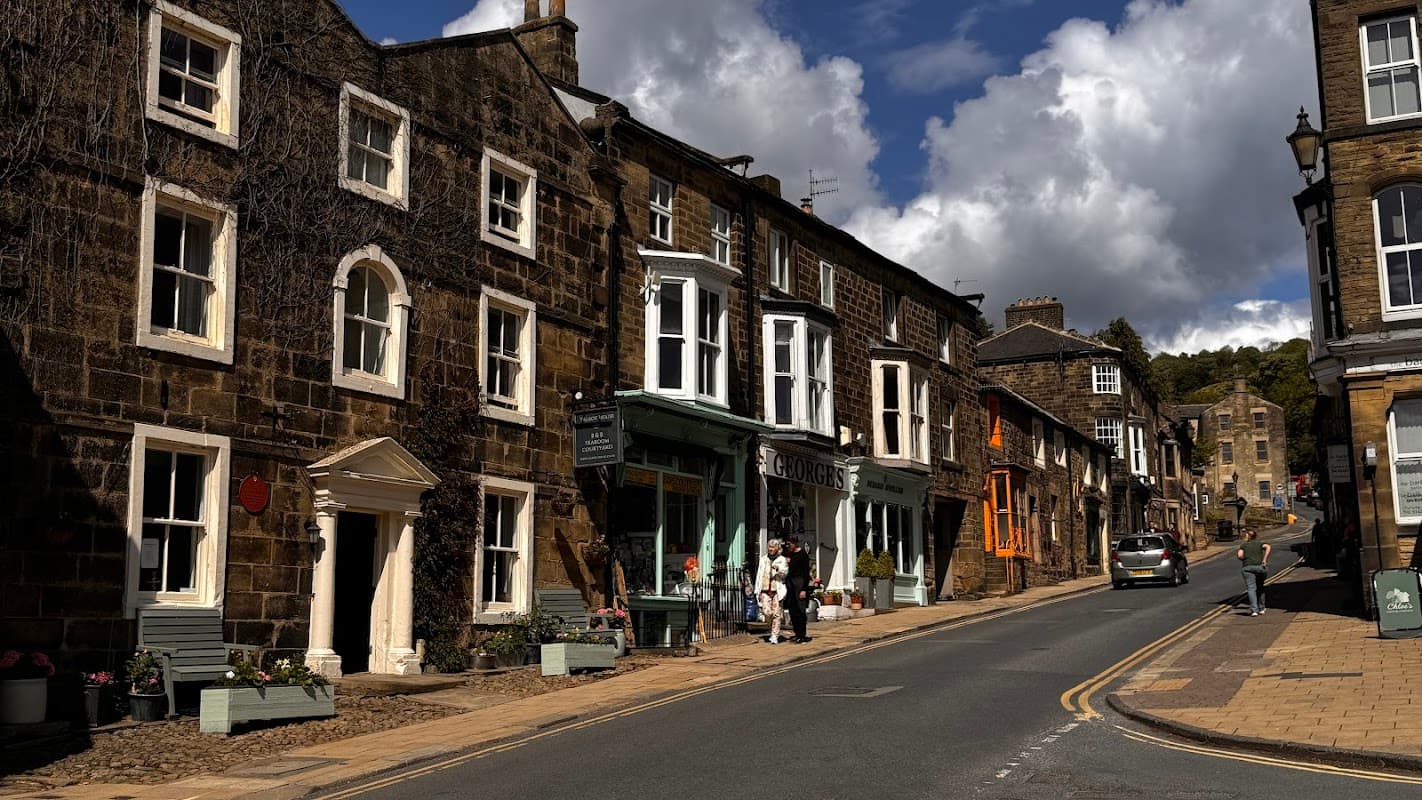 Public car park with stone buildings, shops, and a street leading into the distance under a partly cloudy sky.
