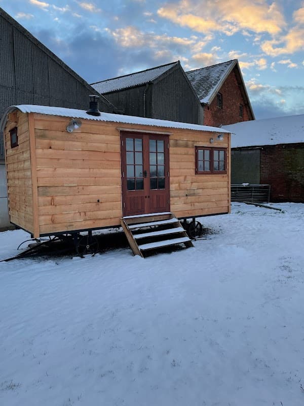 Wooden cabin on wheels in a snowy field, surrounded by farm buildings under a cloudy sky.