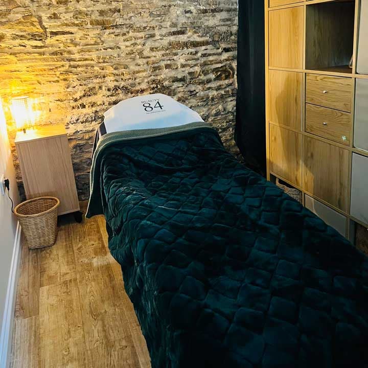 A cozy treatment room featuring a green blanket-covered massage table, wooden storage, and exposed stone walls.