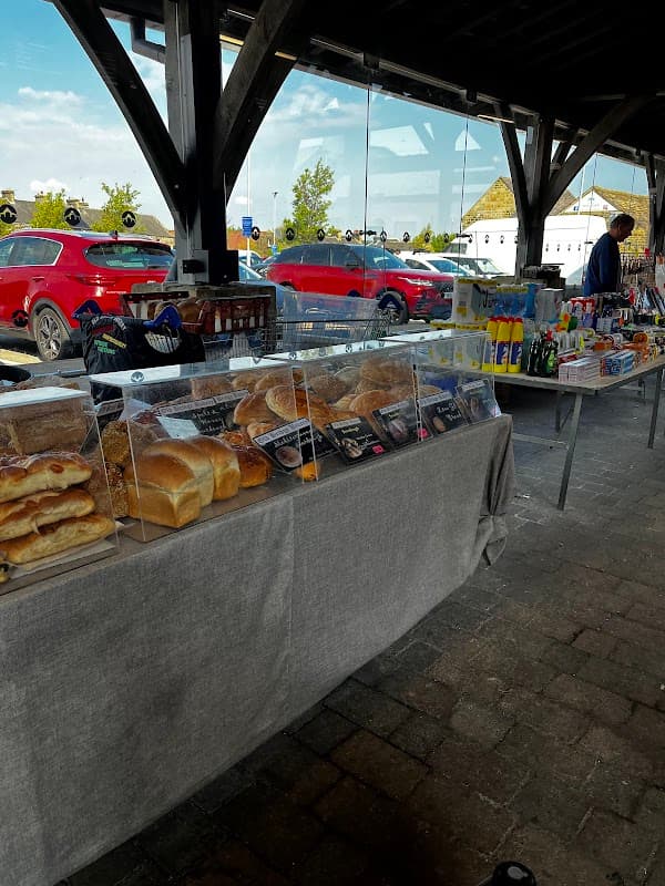 Tables displaying various baked goods and beverages under a wooden structure, with cars parked in the background.