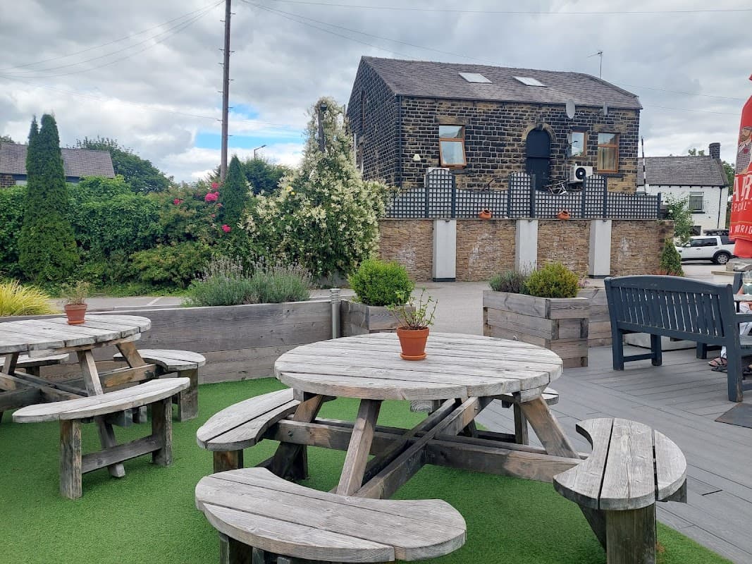 Wooden picnic tables on green grass, surrounded by flowers and shrubs, with a stone building in the background.