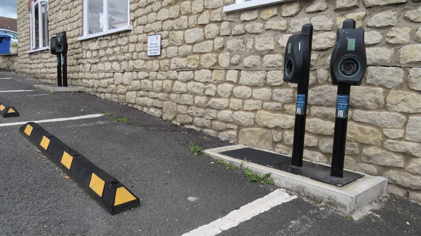Two electric vehicle charging stations mounted on black poles beside a stone wall, with a parking area marked in yellow and black.
