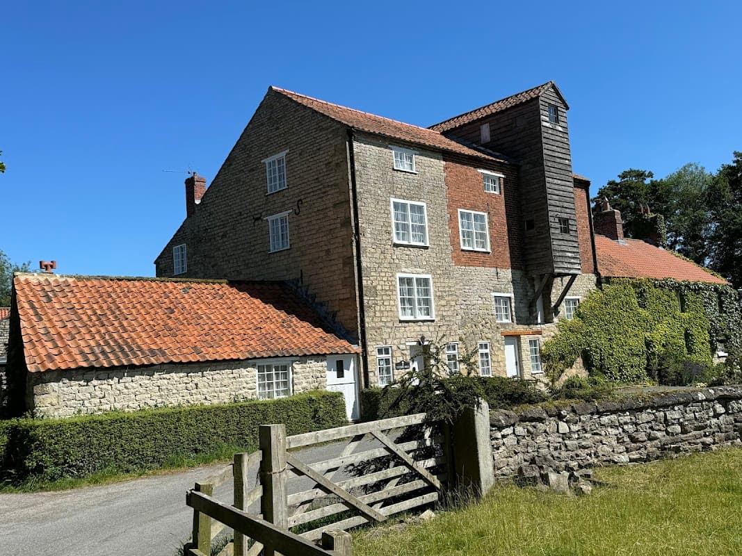 Stone building with red-tiled roofs, surrounded by greenery and a wooden gate, under a clear blue sky.
