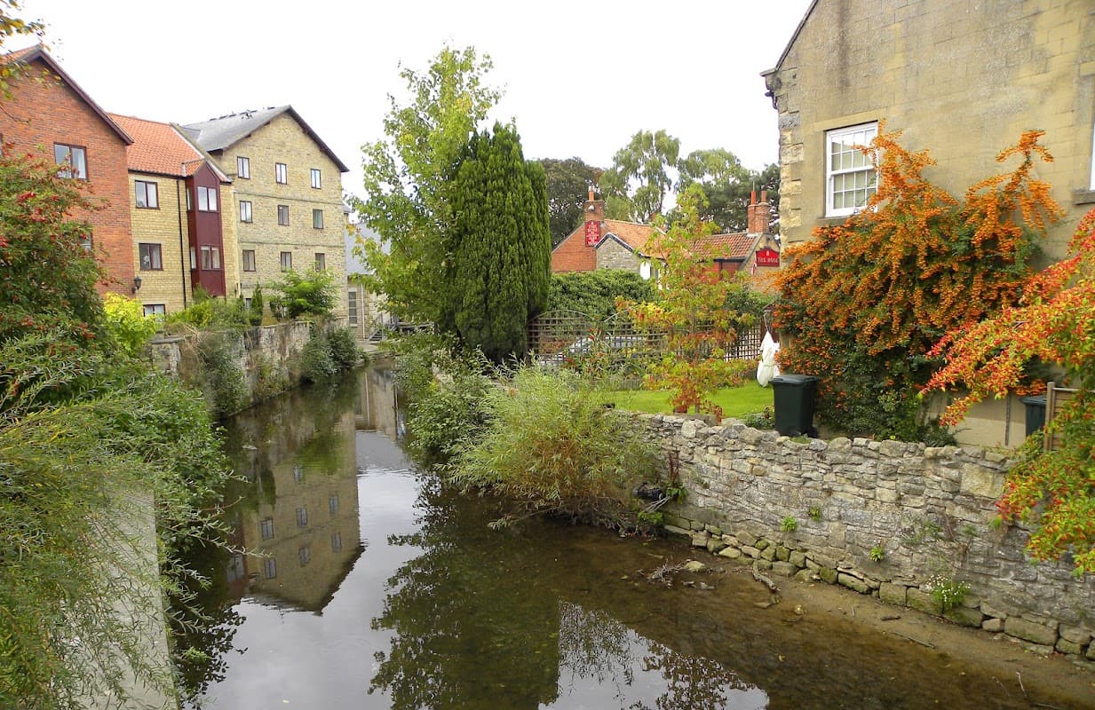 A serene view of a canal lined with stone walls, colorful buildings, and lush greenery in Pickering, Yorkshire.