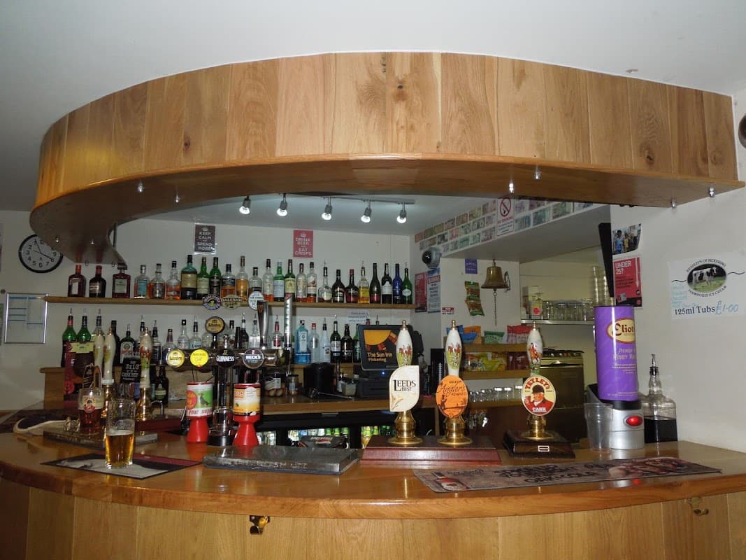 Wooden bar with various bottles and taps, glasses, and a clock on the wall in a cozy pub setting.