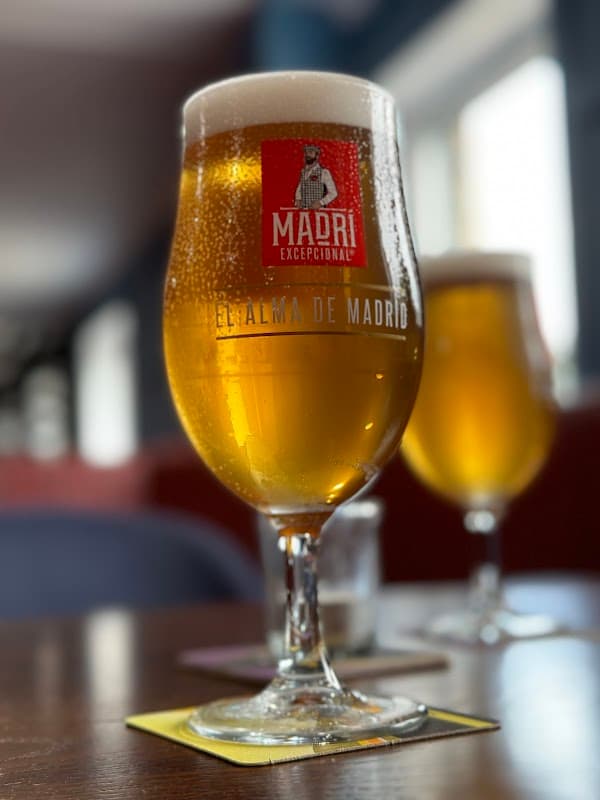 A close-up of a frosty beer glass with a logo, sitting on a wooden table, with a blurred background of another glass.