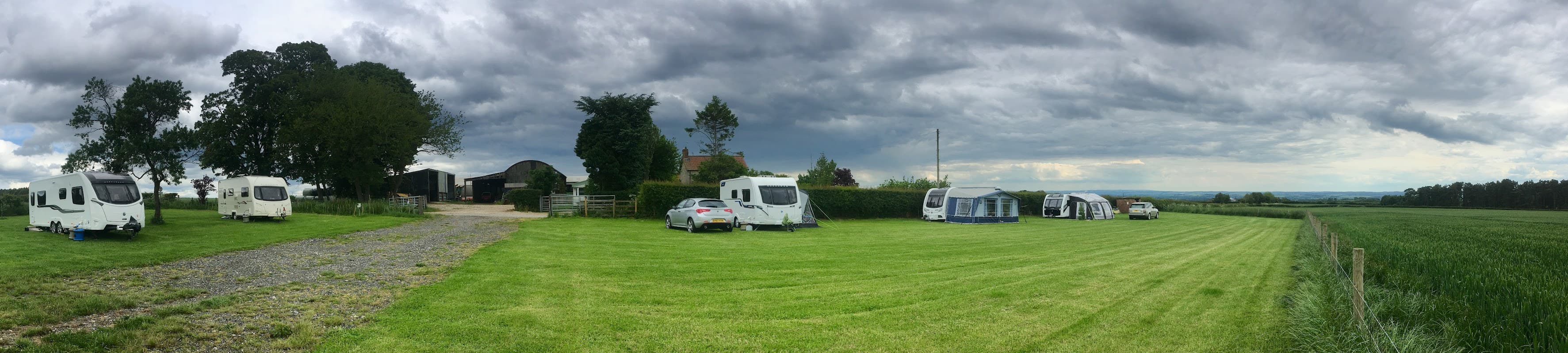 Caravans and tents set in a grassy field under a cloudy sky, with trees and farm buildings in the background.