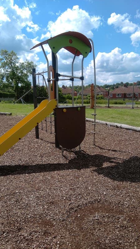 Colorful playground equipment with a slide, set on a gravel surface, surrounded by green grass and houses in the background.