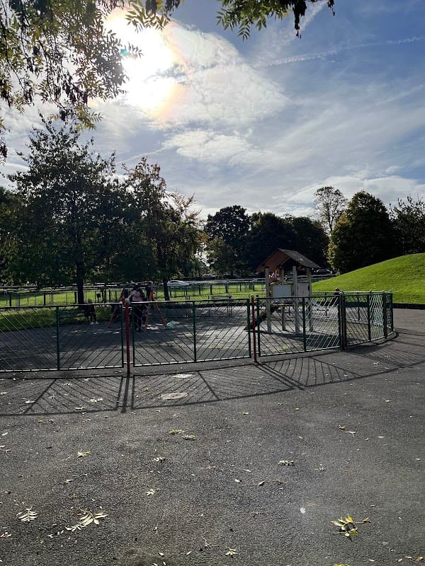 Playground with a fenced area, a play structure, and children playing under a sunny sky with scattered clouds.