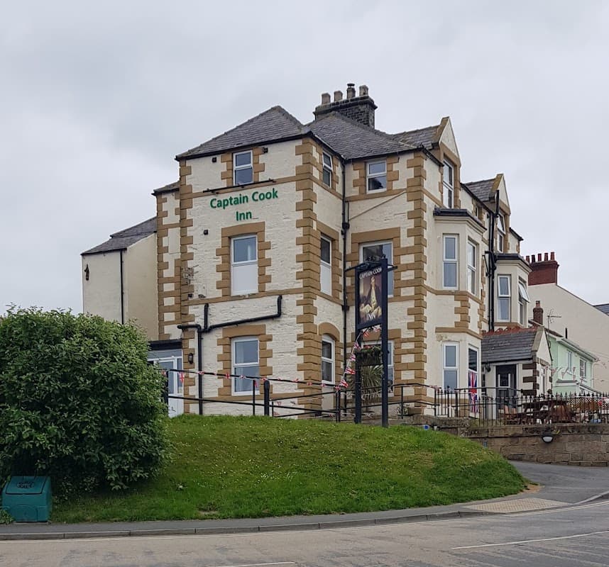Victorian-style building with green signage reading "Captain Cook Inn," surrounded by grass and a cloudy sky.