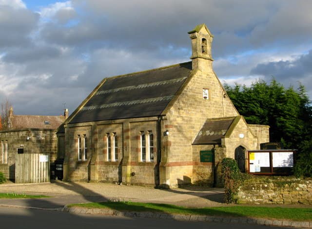 Rainton Village Hall, a stone building with a bell tower, surrounded by greenery and a gravel pathway.
