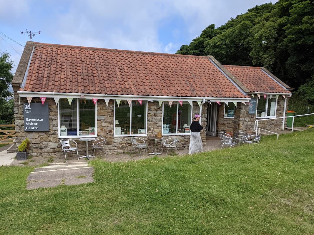Coastal visitor center with stone walls, a red-tiled roof, outdoor seating, and bunting in a green landscape.