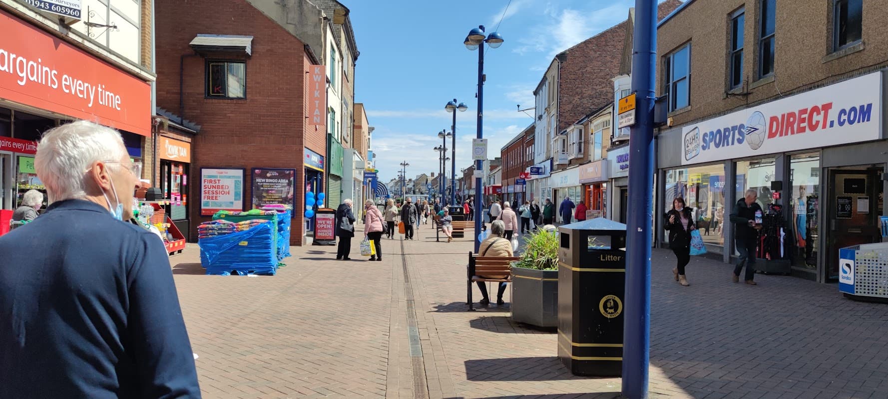 Busy street scene in Redcar with shops, pedestrians, and clear blue skies. Benches and a bus stop are visible.