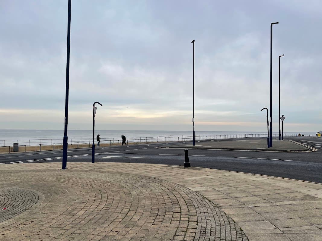View of Redcar High Street East, featuring a wide promenade, streetlamps, and a calm sea under a cloudy sky.