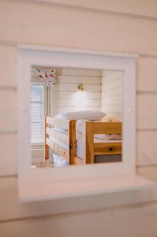 Wooden bunk beds are reflected in a white-framed mirror against a light-colored wall. Natural light enters from a window.