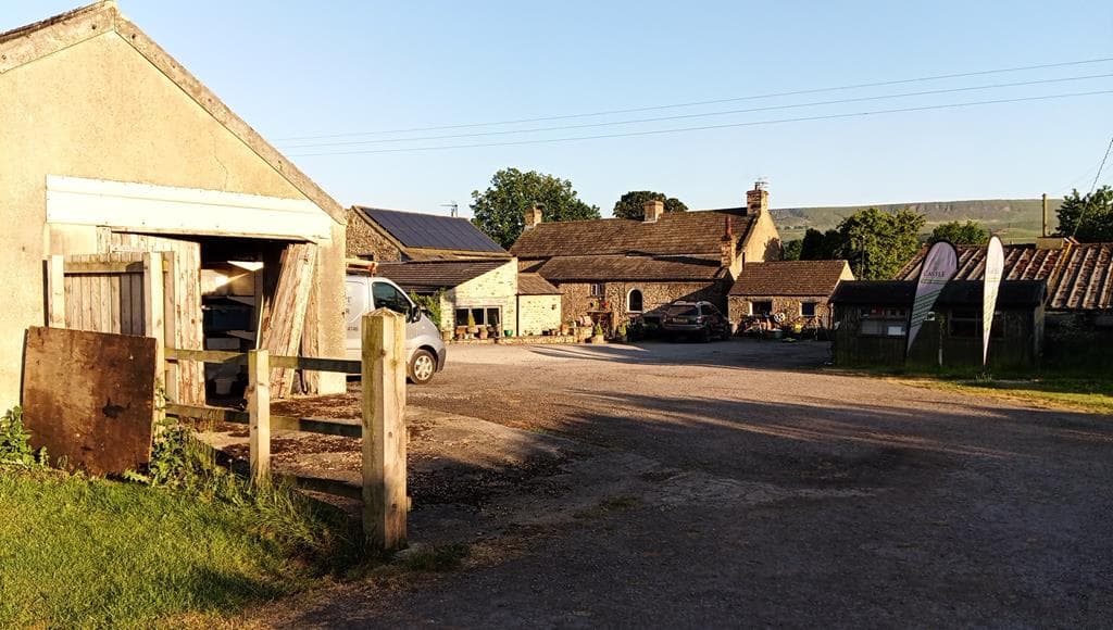A rustic caravan site with stone buildings, parked vehicles, and green fields under a clear blue sky in Redmire, Yorkshire.