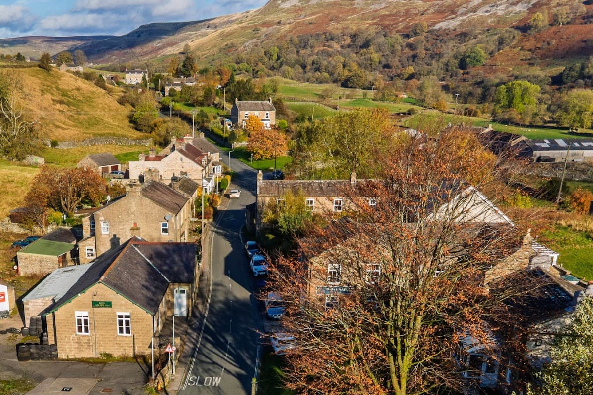 Reeth Memorial Hall nestled among stone buildings, surrounded by lush green hills and autumn-colored trees.