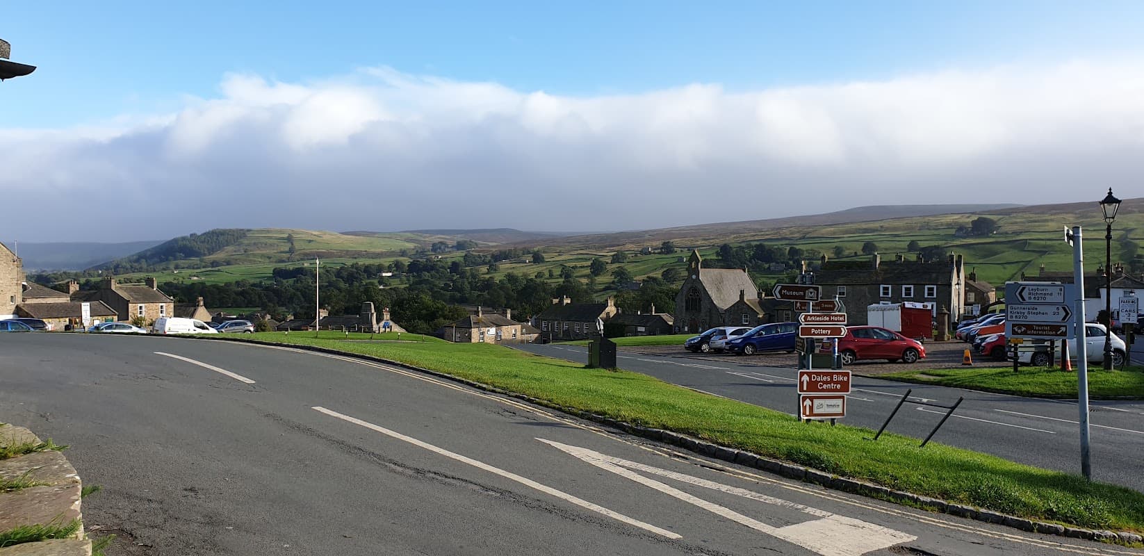 Reeth Public Toilets sign with a scenic view of rolling hills and a village in the background.