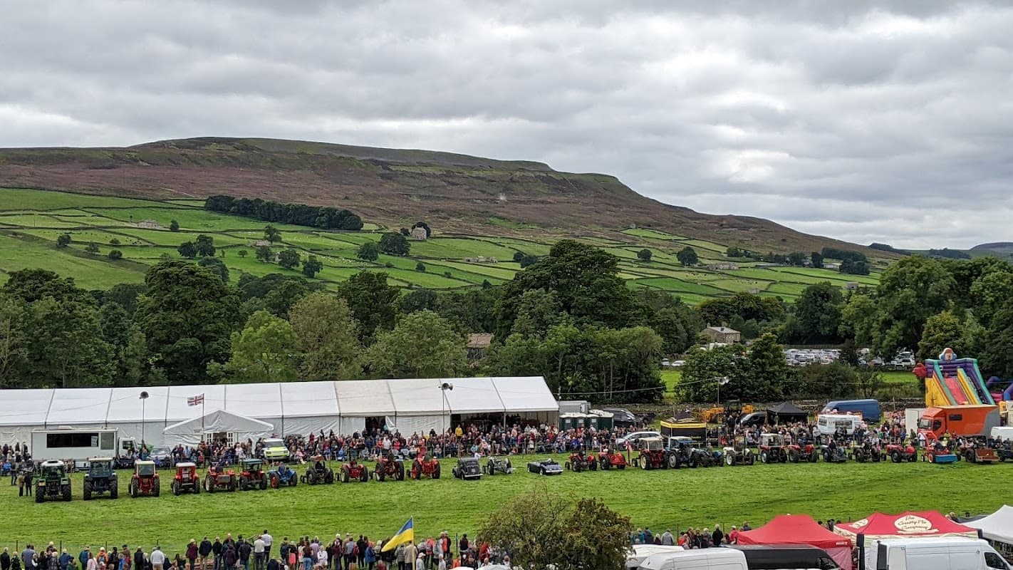 Crowd gathered at the Reeth Show with tractors lined up, lush green hills in the background under a cloudy sky.