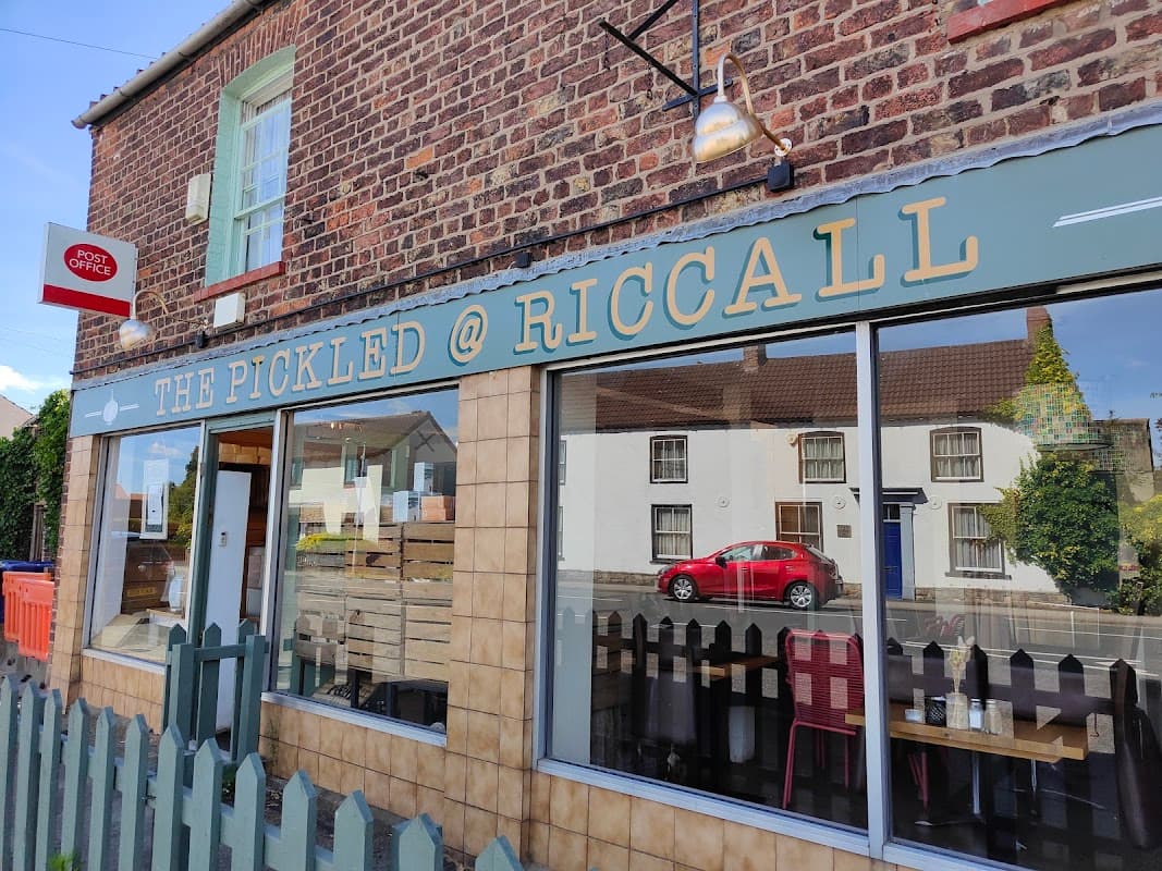 The Pickled @ Riccall sign on a brick building with a red car parked outside and a fence in front.