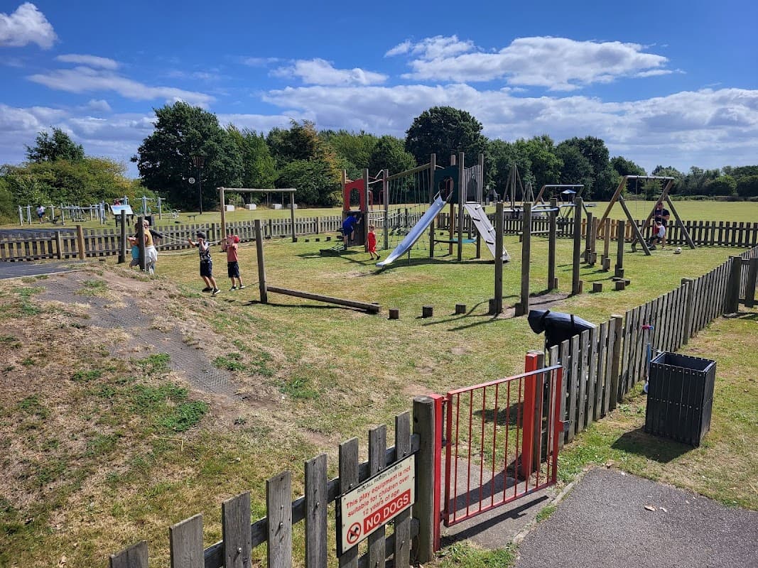 Children playing on various playground equipment in a sunny park with green grass and wooden fencing.