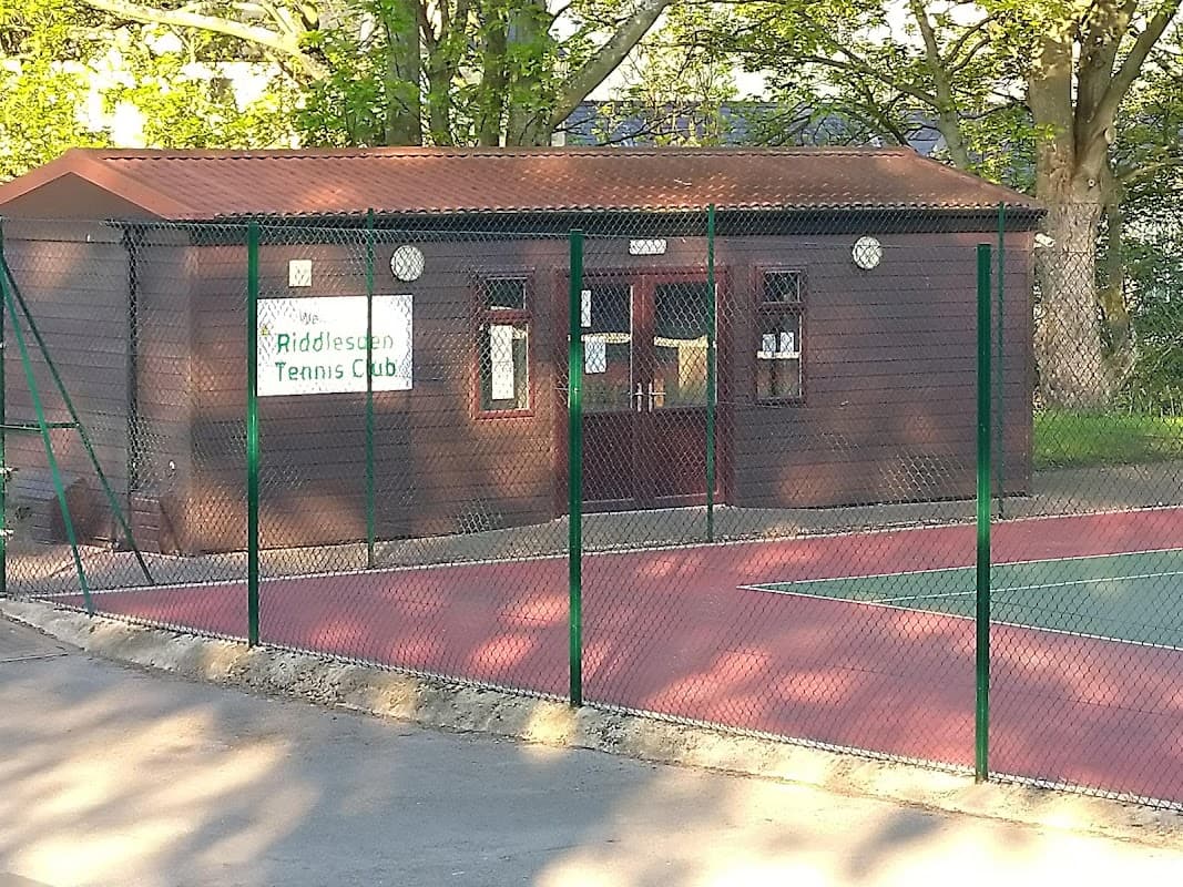 Riddlesden Tennis Club building with a fenced court area and trees in the background.