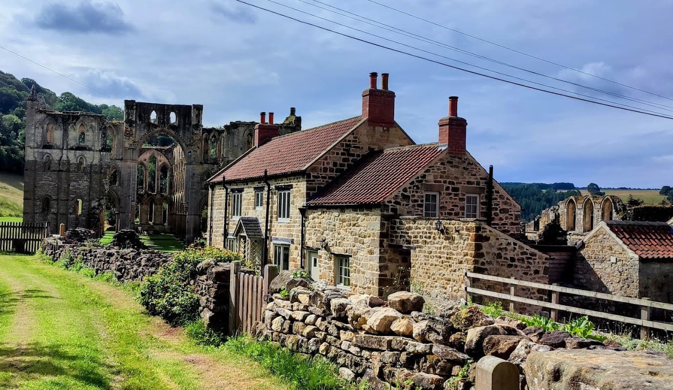 Rievaulx Abbey Visitor Centre with stone buildings, grassy area, and ruins of the abbey in the background.