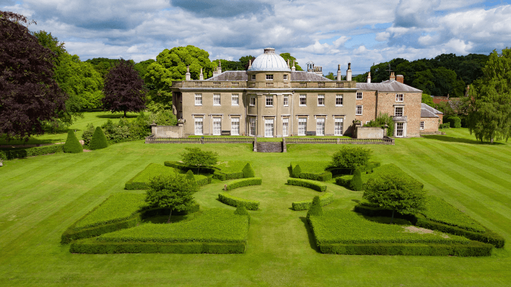 Scampston Hall features a grand building surrounded by neatly trimmed hedges and lush green lawns under a cloudy sky.