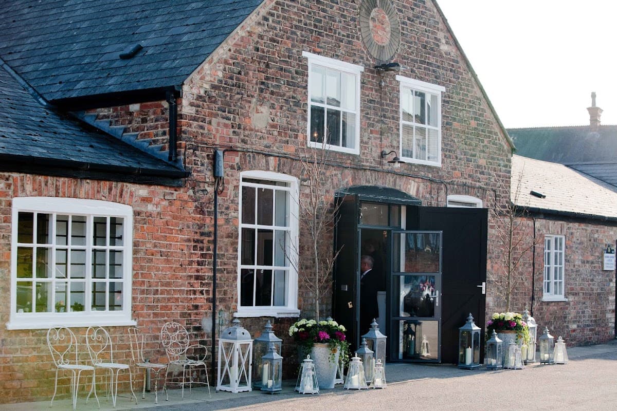 Charming brick building with large windows, black door, and flower arrangements beside lanterns and outdoor seating.