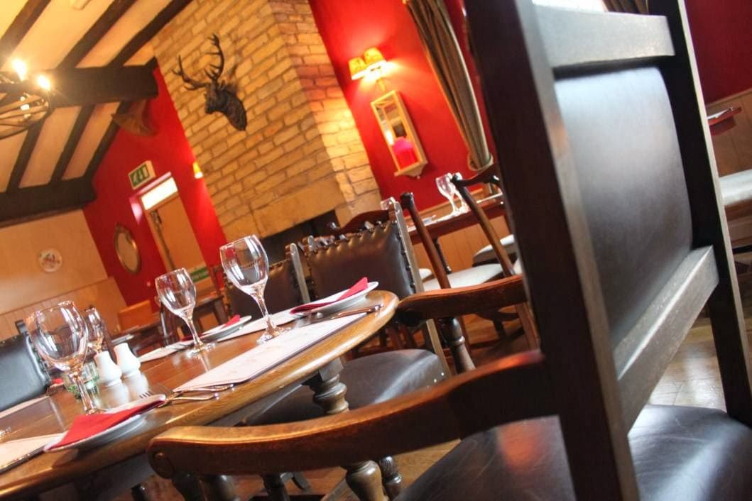 Cozy bar interior with wooden chairs, a stone fireplace, and red walls, featuring a mounted deer head and dining setup.