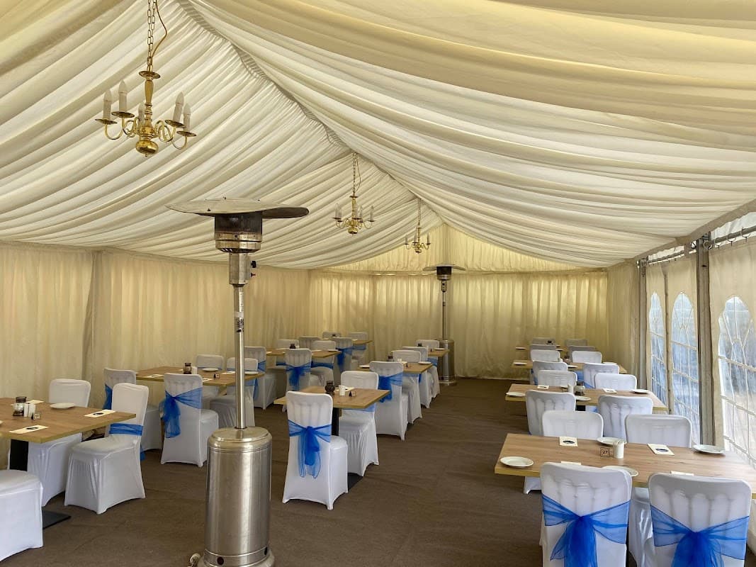 Elegant marquee interior with white chairs adorned with blue sashes, wooden tables, and heating lamps under a draped ceiling.