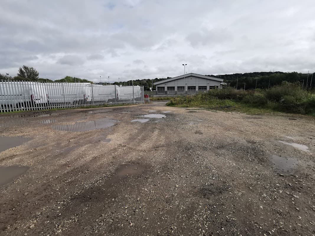 Gravel car park with puddles, surrounded by greenery and a building in the background, near Centenary Riverside Nature Park.