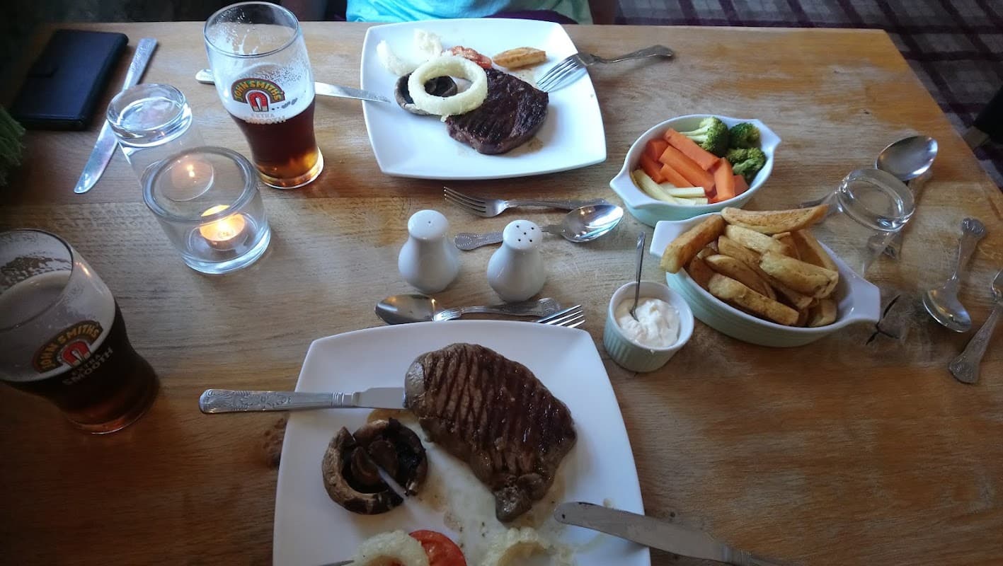Two plates of steak with sides of vegetables, chips, and sauces, alongside two pints of beer on a wooden table.