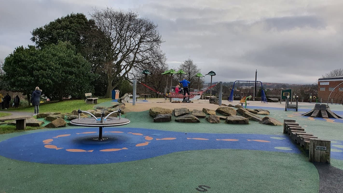 Playground with colorful surfaces, climbing structures, swings, and benches surrounded by trees and rocks in Ruswarp.