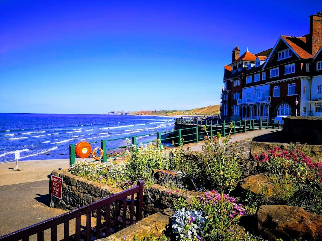 Sandsend beachside view with a car park, vibrant flowers, and a seaside building under a clear blue sky.