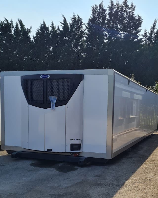 White refrigerated container with a black ventilation grille, parked in a sunny area surrounded by trees.