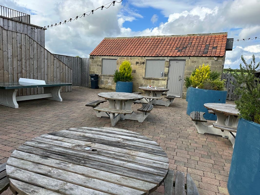 Outdoor seating area with wooden picnic tables, planters, and a rustic building under a cloudy sky.
